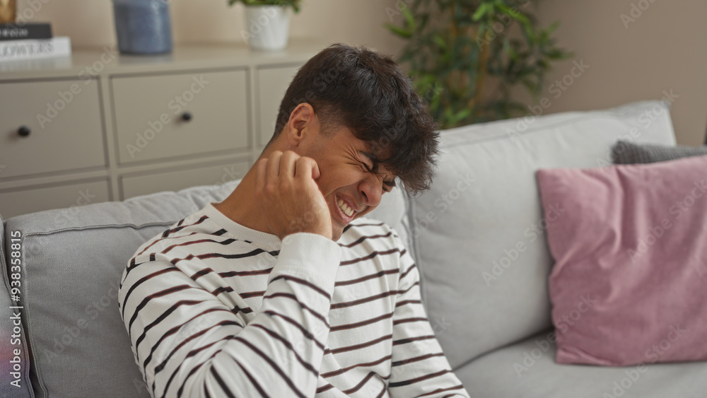 Canvas Prints A young hispanic man scratching his itchy face while sitting on a couch in a cozy living room, displaying an expression of discomfort.