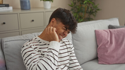 A young hispanic man scratching his itchy face while sitting on a couch in a cozy living room, displaying an expression of discomfort.