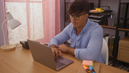 A young hispanic man working on a laptop indoors in an office setting with shelves and various office supplies around.