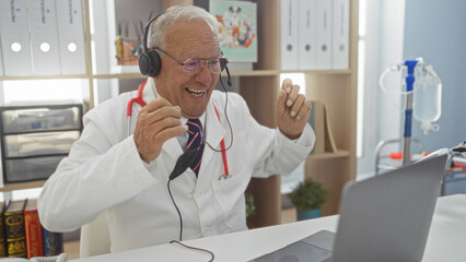 Elderly, grey-haired man in a clinic wearing headphones and a white coat smiles while interacting online in a medical setting.