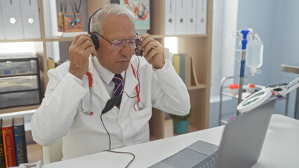 Elderly caucasian man in a clinic wearing a white coat, using headphones, and working on a laptop in a medical office setting.