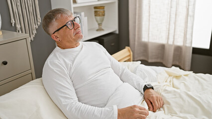 Fototapeta premium Middle-aged man with glasses, wearing a white shirt, relaxing on a bed in a modern bedroom.