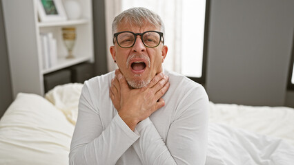 A gray-haired man in glasses expressing throat pain while seated on a bed in a bedroom setting.