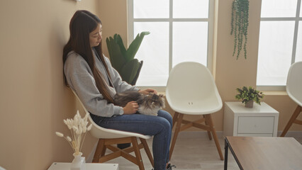 A young chinese woman sits indoors at a veterinary clinic, gently holding her pet cat in the waiting room, surrounded by indoor furniture and plants.