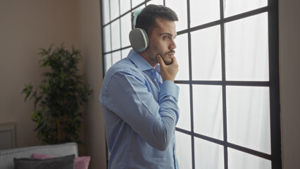 A young hispanic man with a beard wearing headphones stands in a living room, thoughtfully gazing out the large windows of his apartment.