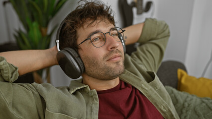 A relaxed young hispanic man enjoys music on headphones in a cozy living room setting, exuding casual comfort.