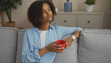A beautiful young african american woman with curly hair sits in a cozy living room, holding a red...
