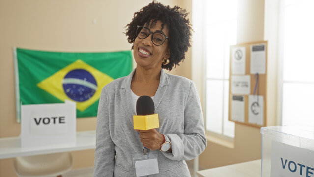 A young, beautiful african american woman with curly hair smiling indoors holding a microphone, standing at an electoral college room with a brazilian flag in the background.