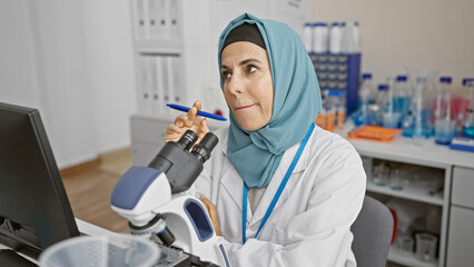 A mature woman scientist in a hijab thoughtfully examines a sample with a microscope in a laboratory setting.