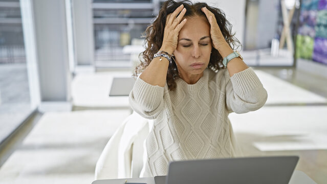 A stressed hispanic middle-aged woman suffering a headache while working in a bright modern office.