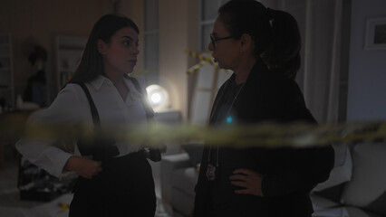 Two women detectives with badges discuss a case at a dimly lit indoor crime scene cordoned off by...