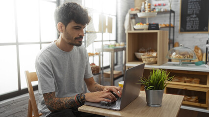 Fototapeta premium Handsome young hispanic man with a beard using a laptop in a cozy bakery interior featuring shelves of pastries and a potted plant on the table
