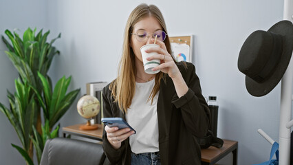 A young woman sips coffee while checking her smartphone in a casually professional office environment, embodying modern work life.