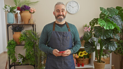 A smiling bearded man in an apron stands in a flower shop filled with various plants, portraying a friendly florist.