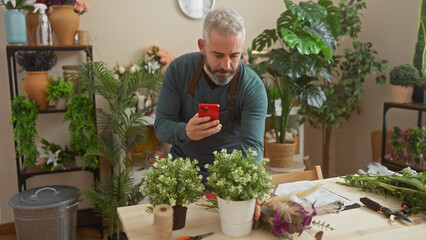 Mature bearded man using smartphone in flower shop surrounded by plants and florist tools.