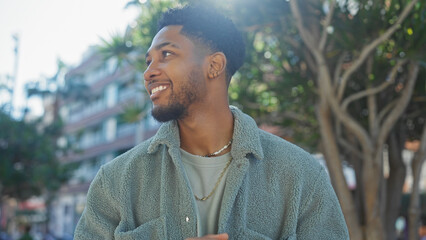 Handsome african american man smiling outdoors in a stylish urban setting with buildings and trees in the background.