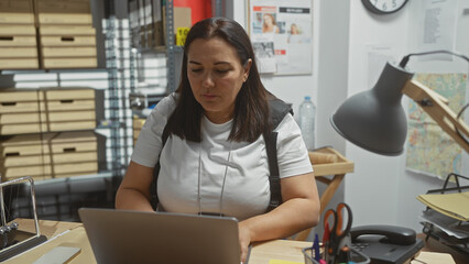 Hispanic woman works at laptop in cluttered police department office, surrounded by files and...
