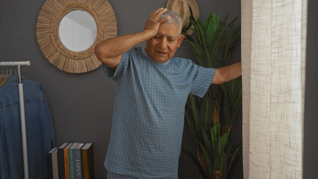 Grey-haired hispanic man feeling dizzy and leaning against the wall in a cozy living room.