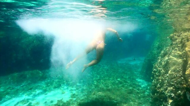 Underwater video. Young girl jumping off a cliff into a blue clear sea. Crystal clear water splashing as she dives off a rough rocky shore underwater, you can see her swimming underwater