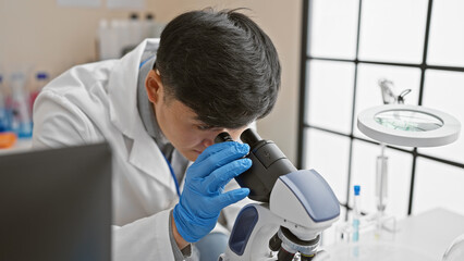 A focused young asian man wearing lab coat and gloves using a microscope in a laboratory setting.