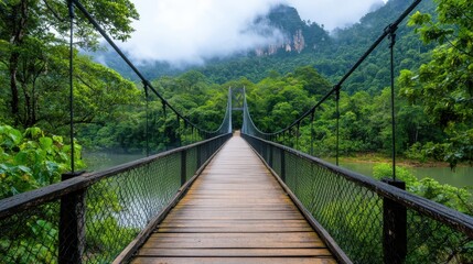 Obraz premium This image showcases a wooden suspension bridge that stretches across a lush green forest, with misty mountains rising in the background, creating a serene and picturesque scene.