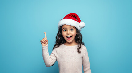 Excited little hispanic girl in santa hat pointing up against blue background. 