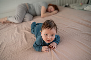 A three-month-old boy lies on his stomach on the bed and his mother sleeps behind him. Postpartum...