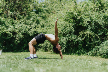 Strong, fit male athlete performing a back bend exercise outdoors in the park on a sunny day,...