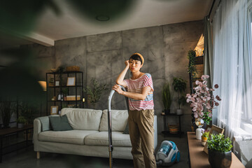 Exhausted, tired mature japanese woman stand with a vacuum cleaner