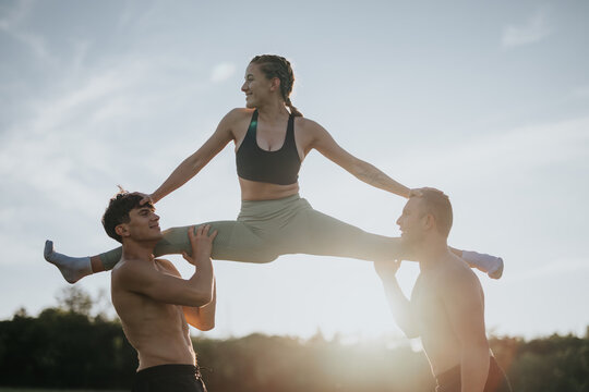 A flexible woman practicing a front split exercise on the shoulders of two male friends against a scenic outdoor backdrop during sunset.