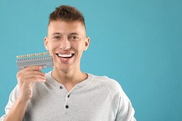 Happy young man with teeth color samples on light blue background, space for text. Dental veneers