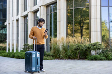 Smiling business traveler using smartphone while pulling suitcase in urban setting. Person wearing orange sweater walking outside office building. Captures concept of business travel © Liubomir