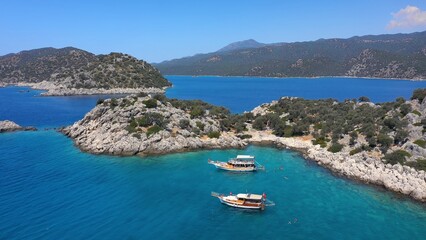 Crystal clear turquoise waters, rugged rocky shores, boats anchored near the coastline, and swimmers enjoying the serene environment. Aerial view of Aquarium Bay at Kekova, Turkey