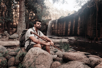 Man with hiking stick next to waterfall in forest.