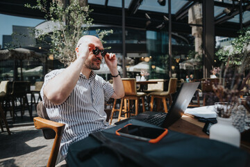 Businessman at an outdoor cafe working on a laptop, adjusting his sunglasses, participating in a business meeting, and enjoying the sunny day.