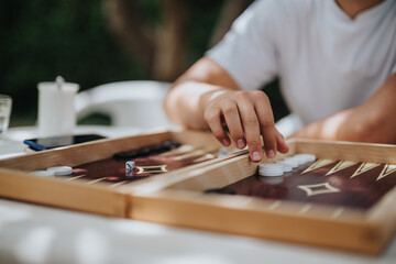 Close-up of a person enjoying a game of backgammon outdoors on a sunny day. The focus is on the hand and game pieces, creating a relaxed and strategic atmosphere.