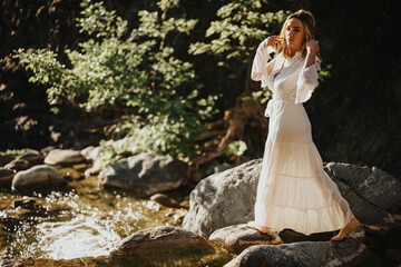 Woman in white dress standing on rocks beside a stream, with sunlight filtering through trees creating a serene natural setting