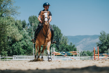 Young rider training on a brown horse in an outdoor arena on a sunny day, with a scenic mountain backdrop.