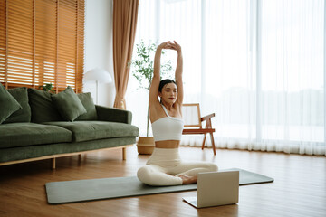 Happy young Asian woman practicing yoga and shoulder stretching at home sitting on floor in living room