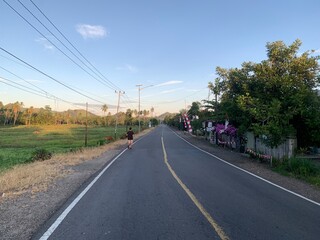 Man Running on a Rural Road at Dawn. A man runs on a straight rural road at sunrise in a tropical country. The road is surrounded by a green field, trees and houses. 
