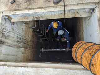 Workers Inspecting a Tank. Two workers wearing safety gear and hardhats are repairing pipes in a deep trench, showing a common scene of industrial work and maintenance.