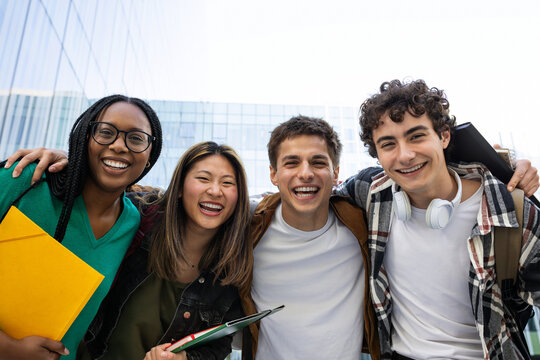 Happy college students embracing, laughing and having fun after class in front of university building looking at camera.