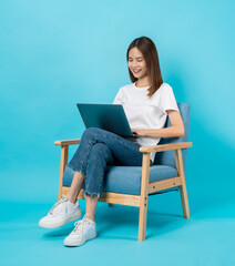 Happy cheerful young woman sitting on the chair and using laptop on blue background.