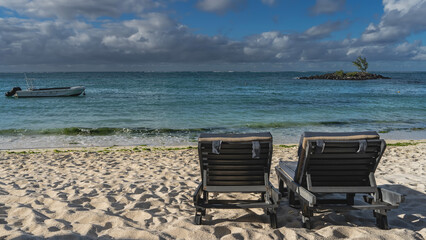 Two sun loungers with soft mattresses stand on  a sandy beach. The motorboat is anchored in the turquoise ocean. A tiny rocky island and a lonely tree in the distance. Blue sky, clouds. Mauritius. 