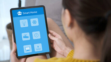 Hand using digital tablet with smart home control icon on screen while sitting on sofa at home,...