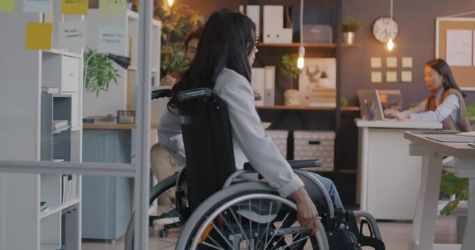 Slow motion back view of handicapped office worker moving in wheelchair to desk ready for work. Inclusive company and disabled employee concept.