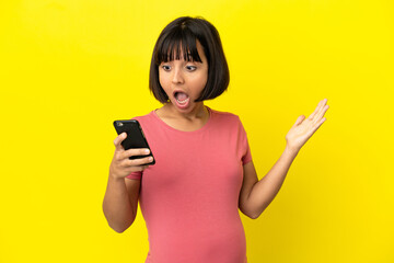 Naklejka premium Young mixed race pregnant woman isolated on yellow background looking at the camera while using the mobile with surprised expression