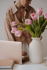 Cozy workspace with pink tulips in a white vase, an open laptop and a stack of notebooks. A person in a beige shirt partially visible in the background, adding a calm, productive vibe