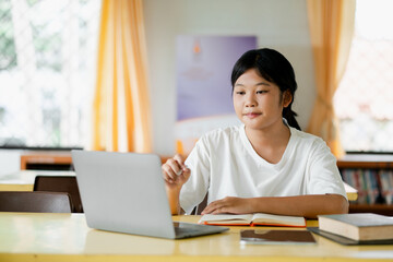 A young student engaged in online learning, using a laptop in a bright classroom with books and notebooks on the desk.