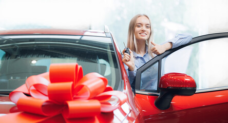 Cheerful Caucasian young woman hold key of vehicle in showroom. Happy owner girl buy first new car...